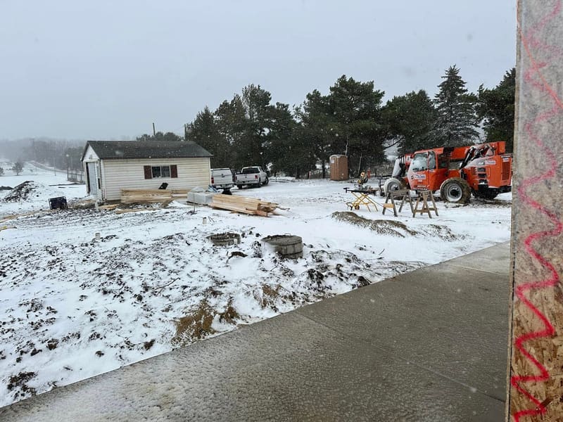 The new clubhouse under construction in the snow during winter 2025, with lumber stacked on the frozen ground and a telehandler parked by the trees