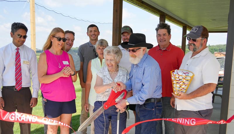 Shirley Maurer cuts the red Grand Opening ribbon with oversized scissors under the pavilion, surrounded by family and Verona community members